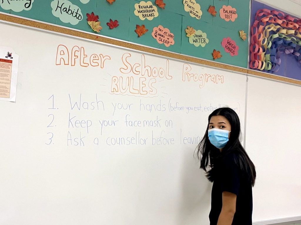 After school program worker in a mask stands in front of a whiteboard that says "After School program rules: Wash your hands before and after you eat and after you enter, keep your face mask on, see a counsellor before leaving"
