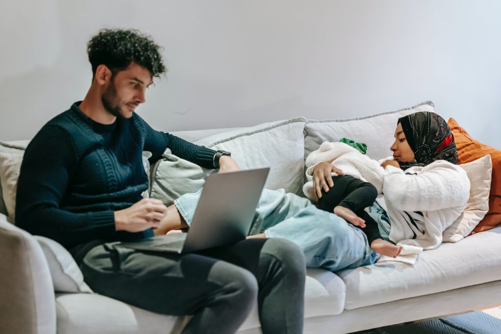 A father sits at his computer with coffee while a mother and child cuddle on the couch beside him